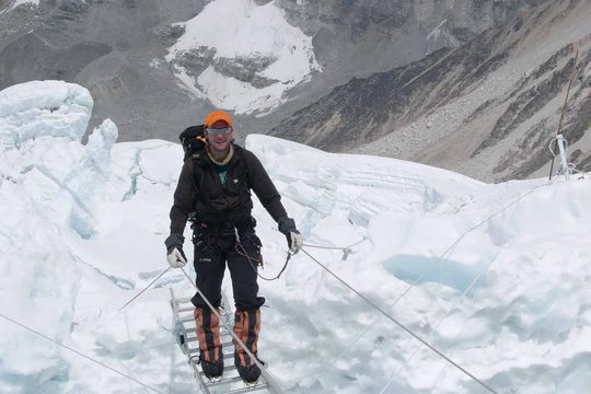 A man wearing thick black clothing and an orange cap hikes through thick snow in the mountains.