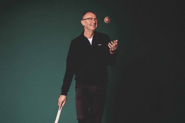 A man wearing glasses and a black jumper stands against a dark green background. He is throwing a cricket ball with one hand.