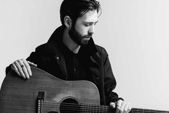 Black and white image of a man in a dark jacket holding a guitar.