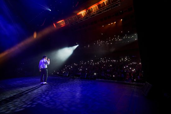 A man stands alone on a stage singing into a microphone. He is lit by a single spotlight and the audience are holding up lights.