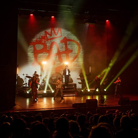 A band perform on a stage lit with yellow strobe lights. Graffiti style text on the back says Bad.
