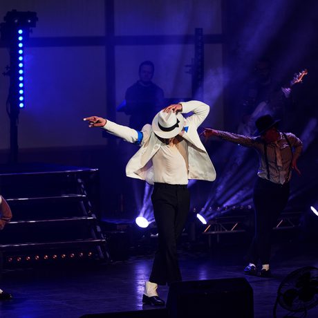 A man in a white jacket and hat dances on a stage lit in blue. He has one arm pointing out to the side and the other holding his hat.