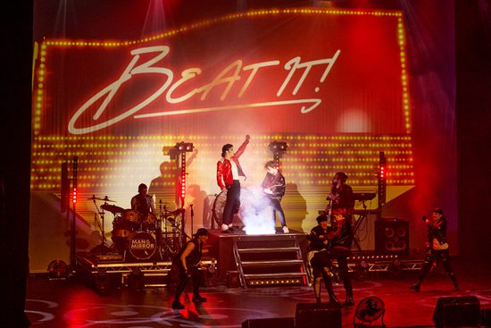 A man in a red leather jacket stands at the top of a small set of stairs on a stage. A band wearing black stand around him. Red neon lights at the back say Beat It!