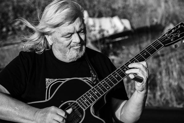 Black and white image of a man with long hair and a beard playing a guitar. he is sat in a grassy field.