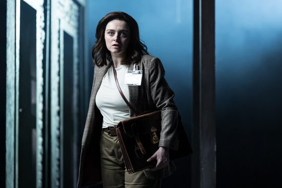A woman in a brown suit holding a brown satchel stands in a doorway, looking scared.
