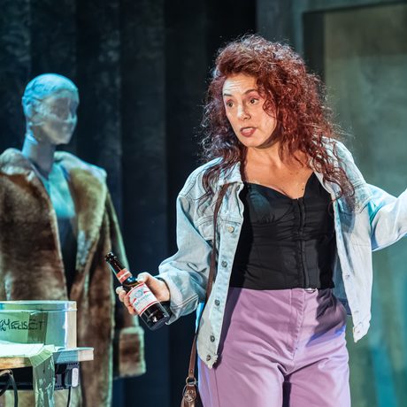 A woman in purple jeans and a denim jacket with red curly hair stands with a beer bottle in her hand. Behind her is a mannequin in a fur coat.