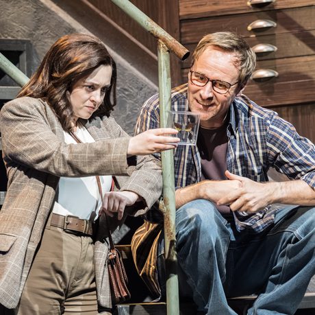 A woman in a brown suit holds up a moth in a case and shows it to a man in a checked shirt sitting on some metal stairs.