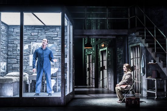 A man in a blue boiler suit stands in a prison cell with glass walls. A woman in a brown suit sits on a chair outside the cell.