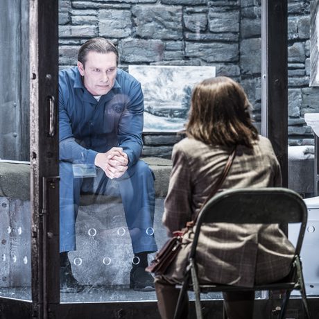 A man in a blue boiler suit sits in a prison cell with glass walls. He looks menacingly towards a woman sat outside the cell.