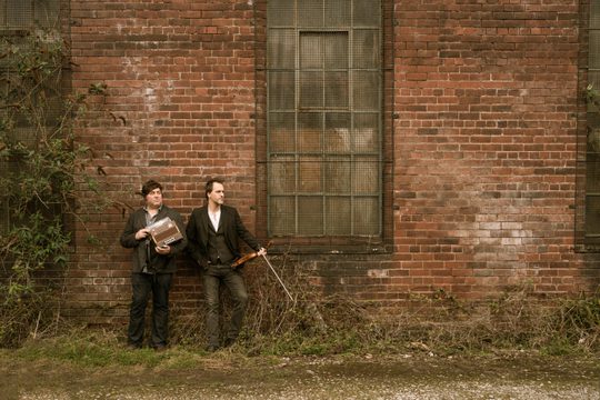 Two men holding instruments stood in front of a derelict red brick building.