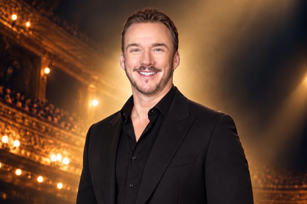 A man in a black shirt and jacket stands in an ornate gold theatre.