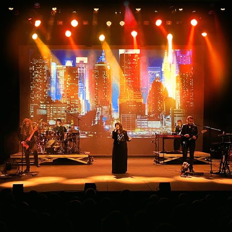 A band performing on a stage. A screen at the back of the stage shows skyscrapers, and they are lit with orange light.