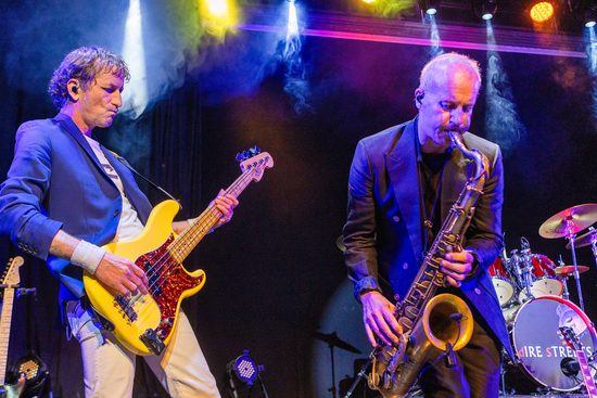 Two men in suit jackets play in a band on stage. One plays a yellow bass guitar and another plays a saxophone.