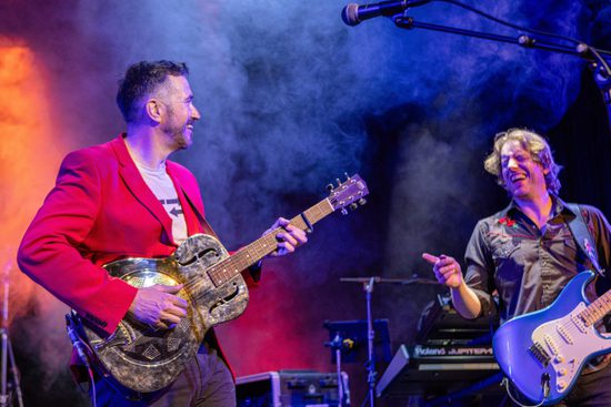 A guitarist in a red jacket and a guitarist in a black shirt laugh together while playing guitars,