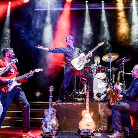A 5-piece band play on stage lit in red, white, and yellow. There are three guitarists, a saxophonist, and a drummer. One guitarist stands at the top of some stairs with his arm out, and the other musicians look at him.