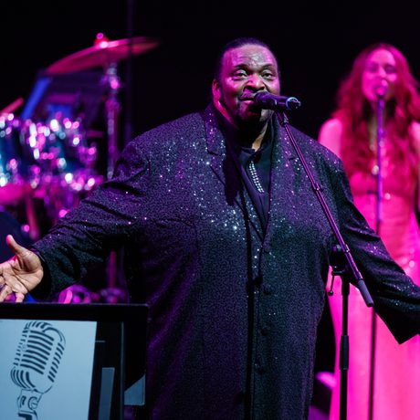 A man in a black sparkly suit stands and sings into a microphone. He holds his hands out to the sides.