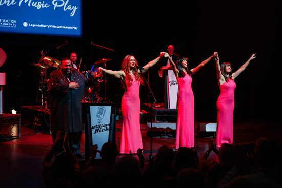 One man in a glittery black shirt and three women in pink dresses stand on stage. They hold hands and bow.