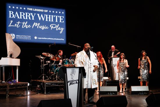 A man in a white suit stands on stage and sings. Three women in silver sparkly dresses and a band perform behind him.
