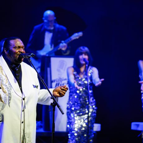 A man in a white sparkly suit stands on stage and sings into a microphone. Three women in silver sparkly dresses stand behind him. They are all lit in blue.