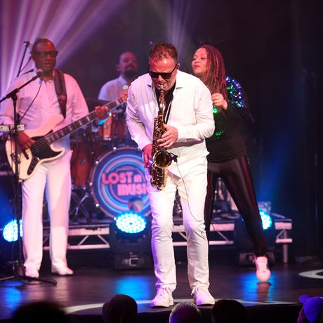 A man in an all-white suit and black sunglasses plays the saxophone.