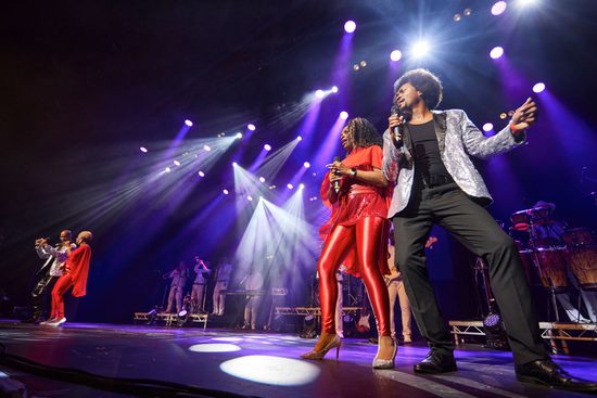 A stage lit with purple lighting. Performers on the stage wearing red, black, and silver sing, accompanied by a live band.