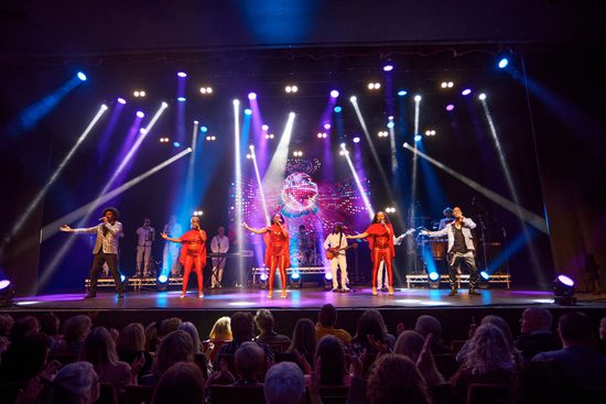 5 singers wearing red, silver, and black sing on a stage with one arm extended to their side. They are performing to an audience, with a live band accompanying them. There are white, purple, and blue spotlights covering the stage.