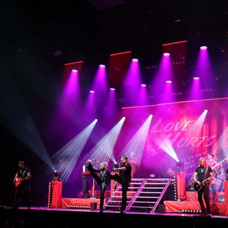 Multiple performers wearing black performing on a stage lit in pink. The two performers at the front are kicking their leg in the air.