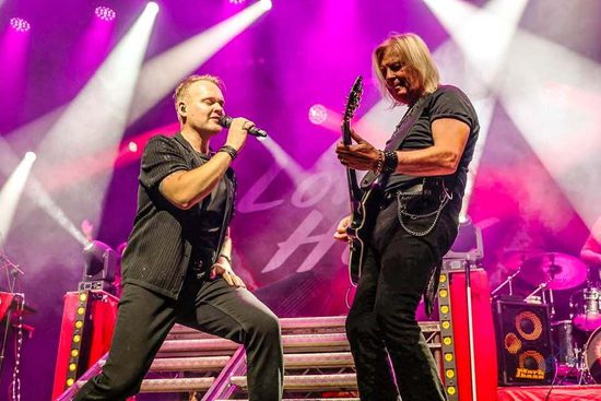 Two performers wearing black stand on a stage lit in pink. One is singing into a microphone, and one is playing the electric guitar.