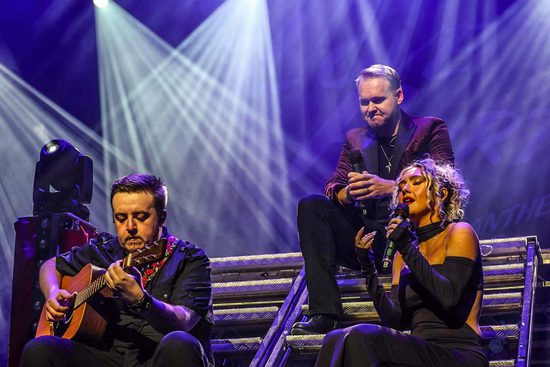 Three performers, two singing and one playing guitar, sitting on metal stairs. They are lit in purple.