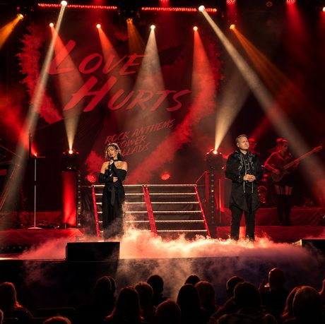 A group of performers wearing black on a stage lit in red. There is smoke covering the floor of the stage.