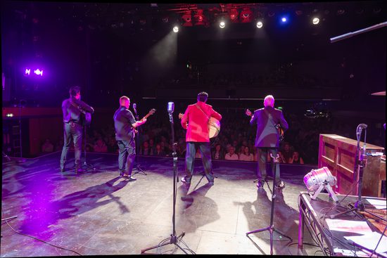 Four musicians photographed from the back, playing instruments to an audience. Three are wearing black and one is wearing a bright red jacket.