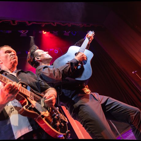 Tow men wearing black stand shoulder-to-shoulder playing guitars. The stage is lit in red.