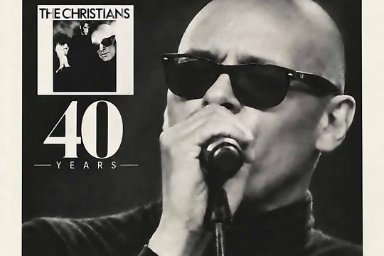 Black and white image of a bald man wearing sunglasses and singing into a microphone.