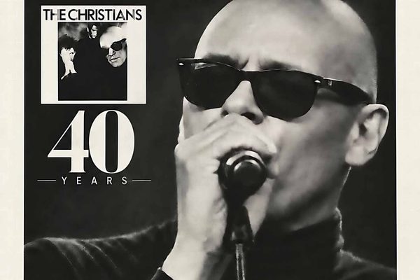 Black and white image of a bald man wearing sunglasses and singing into a microphone.