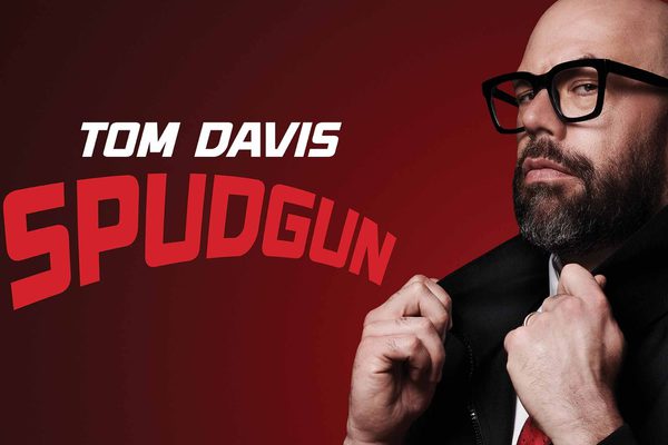 A bald man with a beard wearing a black and white tracksuit and thick glasses stands against a deep red background. Text: Tom Davis - Spudgun.