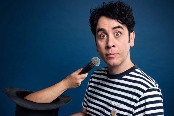 A brunette man in a navy and white stripy top looks alarmed. There is a hand coming out of a black top hat, holding a microphone to his mouth. He is against a dark blue background.