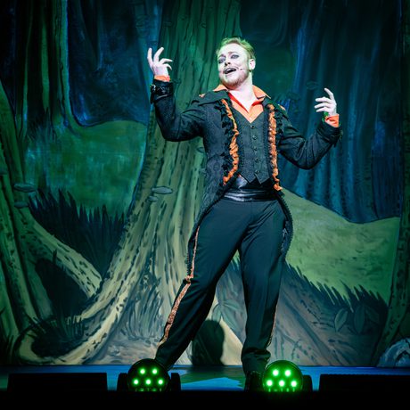 A man in a black suit with orange trim laughs in an evil way.