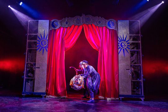 Two puppeteers on the puppet of a tiger in front of red stage curtains
