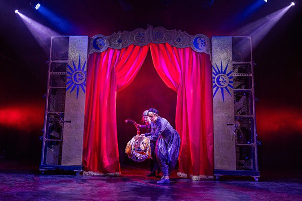 Two puppeteers on the puppet of a tiger in front of red stage curtains