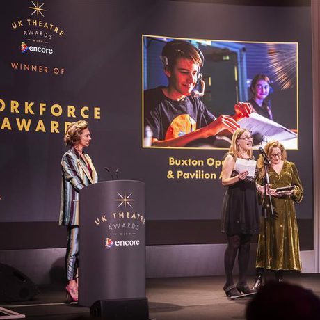 Two members of staff accept and award on stage from a woman in a striped suit.