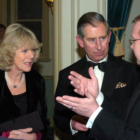 King Charles and Queen Camilla dressed in black tie listening to a man speaking to them