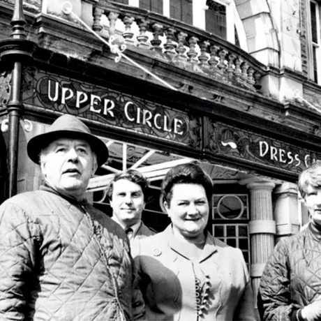 Black and white: % people stand out side the front of the opera house, the balcony above the entrance can be seen above them