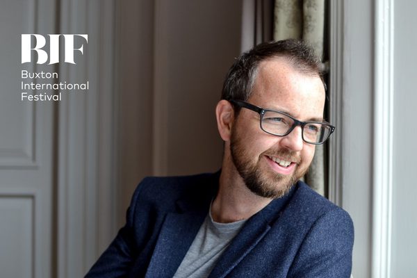 A man with dark, charcoal hair and a trimmed beard is wearing glasses, a grey t-shirt, a blue blazer is propped up against a curtain and staring from out of a window.
