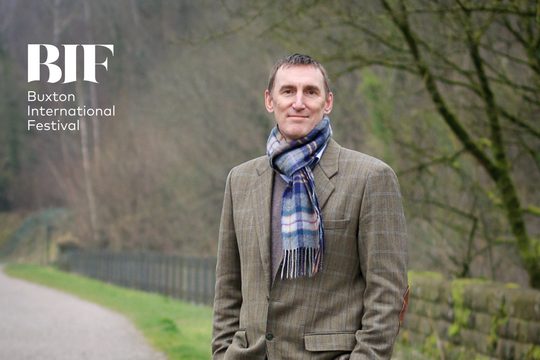 A man with short brown hair, wearing a light brown, grid-patterned blazer and a blue chequered scarf, stood on a path against a backdrop of trees and grass.